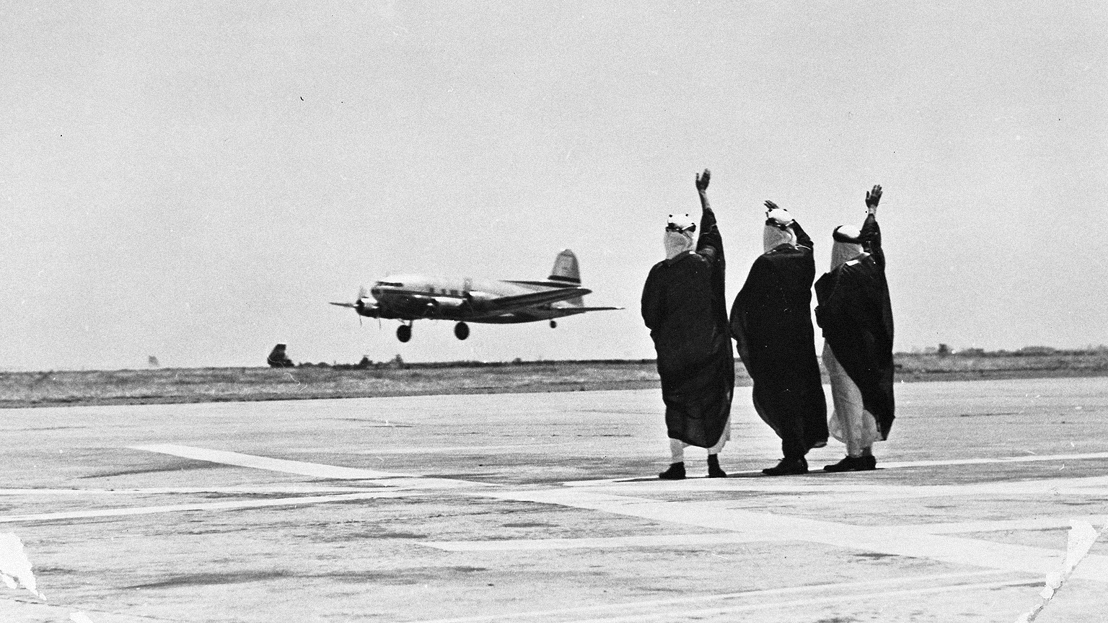 Three people waving at an airplane flying by at a landing strip