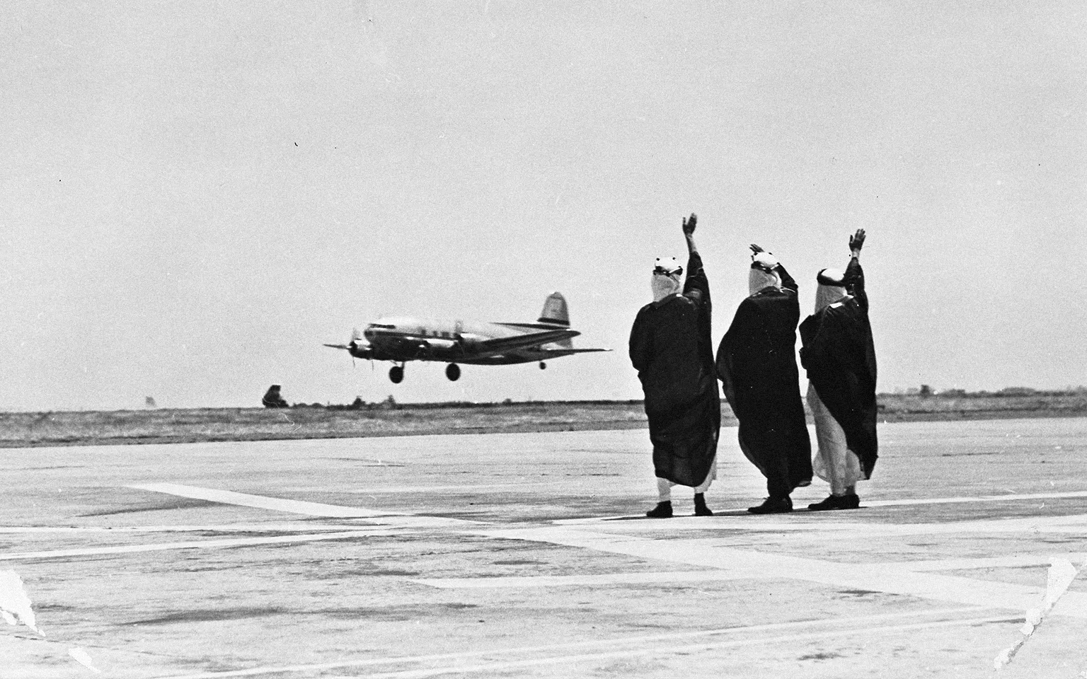 Three people waving at an airplane flying by at a landing strip