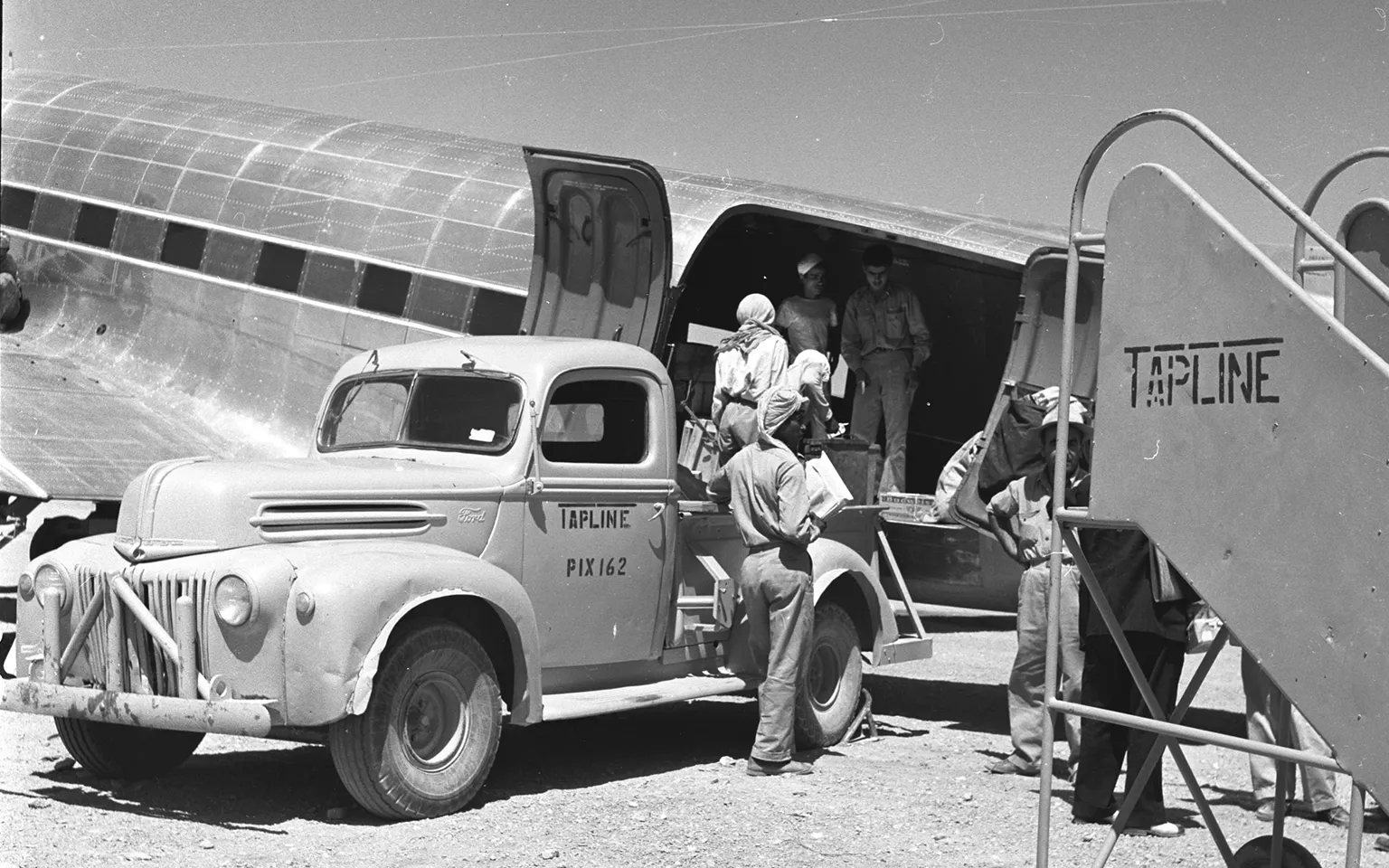 Truck parked in front a Mukamalah airplane and people carrying cargo